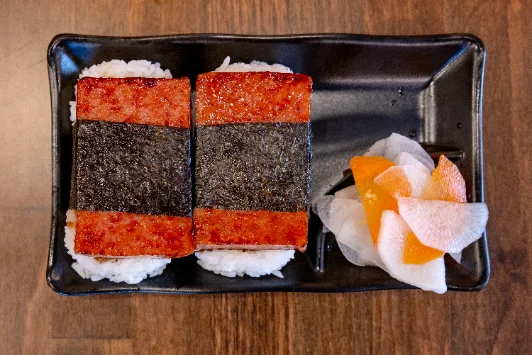 Spam musubi on a black plate with pickles